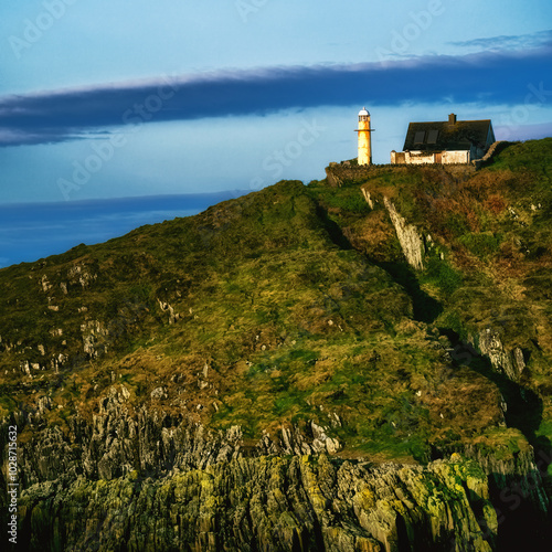 A white lighthouse stands on a rocky cliff, next to a small, white, single-story house with a grey roof. The lighthouse is tall and slender, with a white cylindrical body and a black lantern. photo