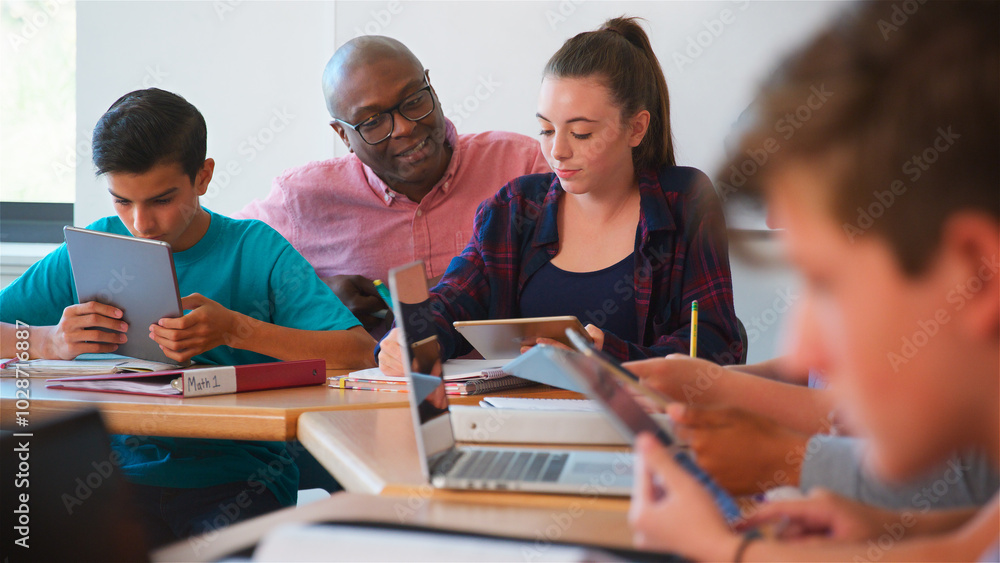 High School Teacher Helping Female Student Sitting At Desk With Digital Tablet In Classroom High School Teacher Helping Female Student Sitting At Desk With Digital Tablet In Classroom