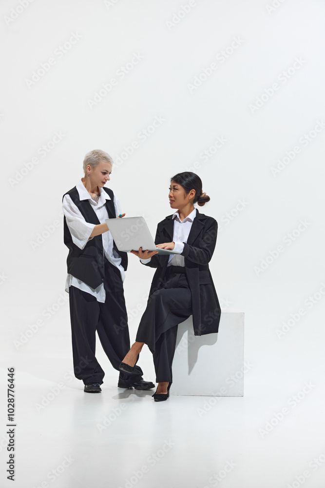 Colleague joins in to discuss something with seated woman, both smiling while engaging over laptop against white studio background. Concept of DEI, business, partnership, work, support and unity.