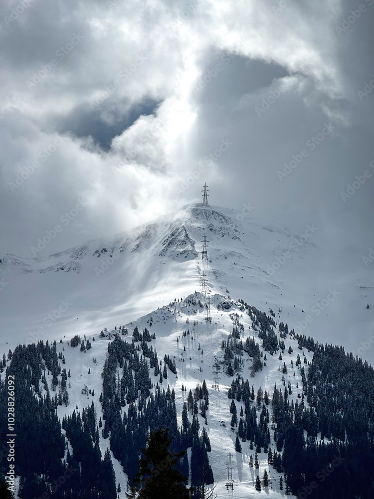 Electric power line runnnig up a snow covered mountain with snow line at the bottom in the ski region of Saalbach-Hinterglemm, Austria against dramatic cloudy sky