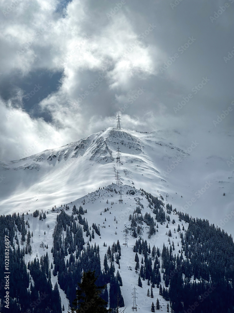 Electric power line runnnig up a snow covered mountain with snow line at the bottom in the ski region of Saalbach-Hinterglemm, Austria against dramatic cloudy sky
