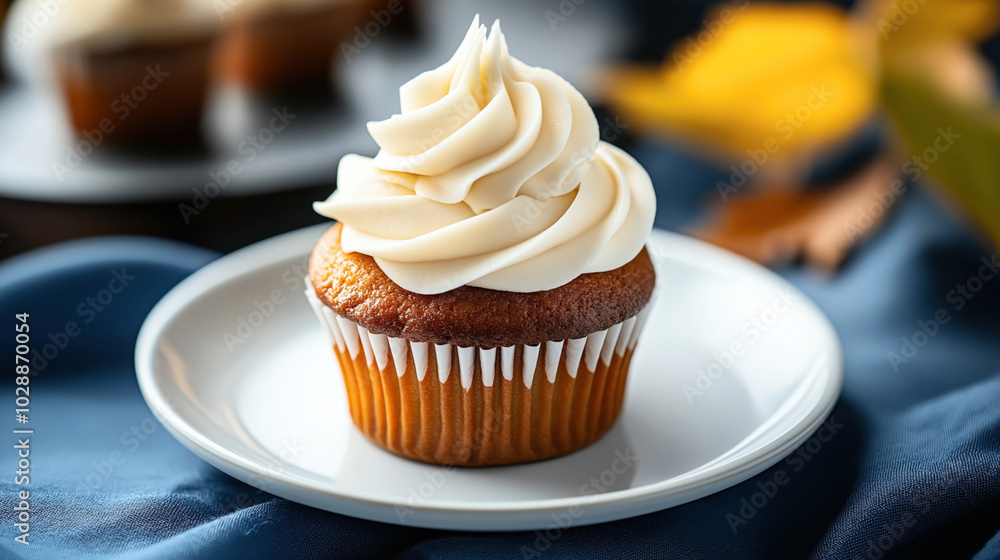 Close-up of a vanilla cupcake with white frosting on a white plate against a blurred background featuring textured fabric and soft lighting. Close-up of a vanilla cupcake with white frosting on a white plate against a blurred background featuring textured fabric and soft lighting.
