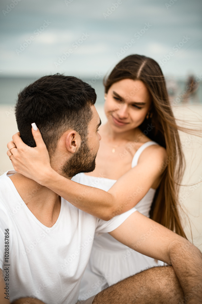 Young girl hugs her husband by the neck and looks into his eyes smiling while sitting on the sand at the beach Young girl hugs her husband by the neck and looks into his eyes smiling while sitting on the sand at the beach