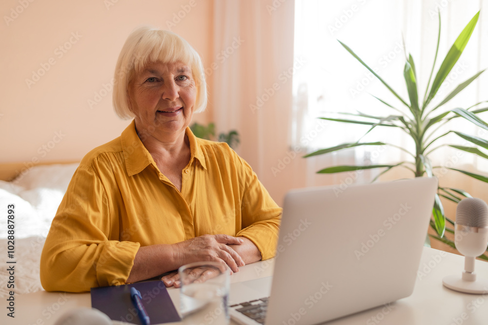 Happy senior woman looking at camera while working at home with laptop. Beautiful stylish elderly lady browsing site on cellphone. High quality photo