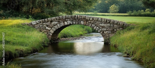 A stone arch bridge over a meandering stream, surrounded by lush green grass and trees.