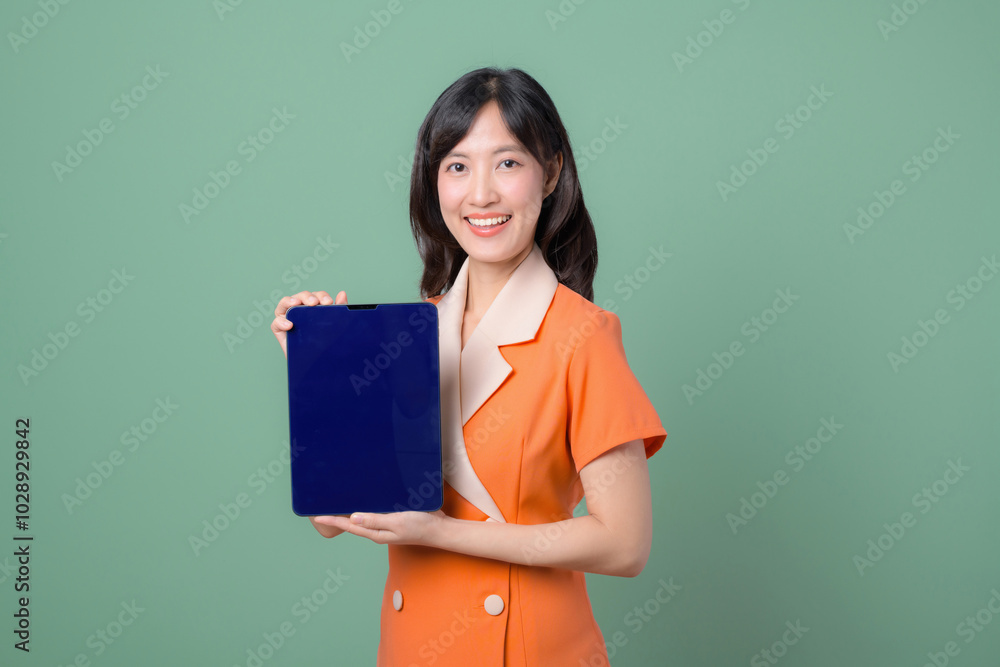 A young woman in an orange dress with white accents stands against a green background, holding up a blank tablet with both hands. She smiles warmly at the camera. A young woman in an orange dress with white accents stands against a green background, holding up a blank tablet with both hands. She smiles warmly at the camera.