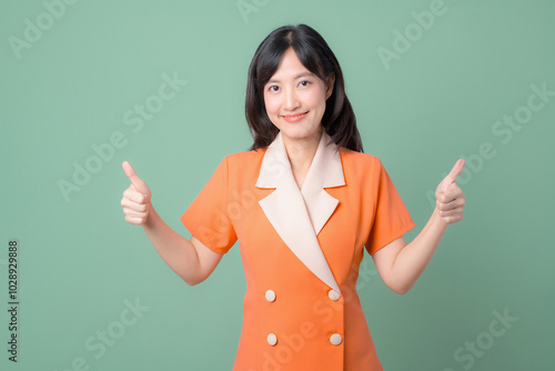 A woman in an orange dress showing double thumbs up with a smile against a green background.