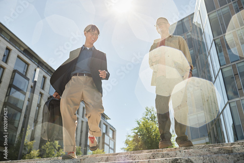 Low angle shot of two smiling male colleagues of different ethnic background walking down stairs enjoying talk and sunny day on way to work in city street, boleh effect photo