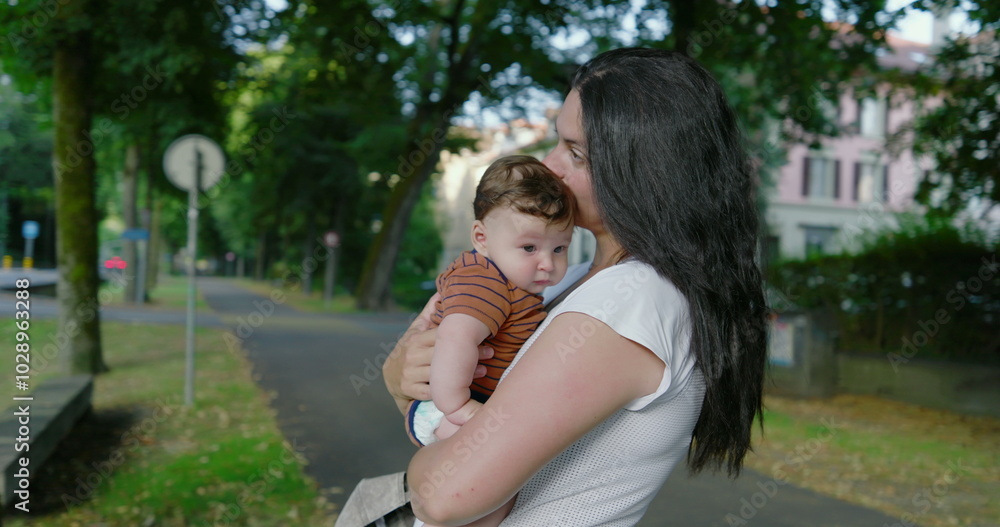 Fototapeta premium Mother holding her baby on a tree-lined urban path, gazing lovingly at her child, surrounded by buildings and greenery, with a calm and nurturing atmosphere captured