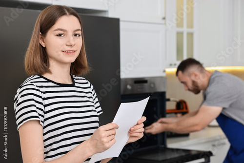 Smiling woman with sheet of paper and repairman fixing oven in kitchen, selective focus