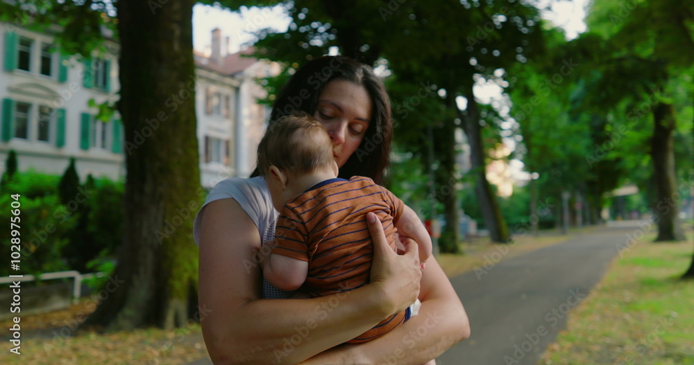 Fototapeta premium Mother cradling her baby close while standing on a tree-lined path, showing deep affection and love, with urban buildings and trees in the background