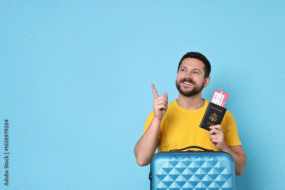 Happy man with suitcase, passport and ticket pointing at something on light blue background, space for text Happy man with suitcase, passport and ticket pointing at something on light blue background, space for text