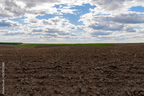 A wide plowed agricultural field extends towards the horizon, showcasing rich brown soil under a sky filled with fluffy clouds and patches of green in the background