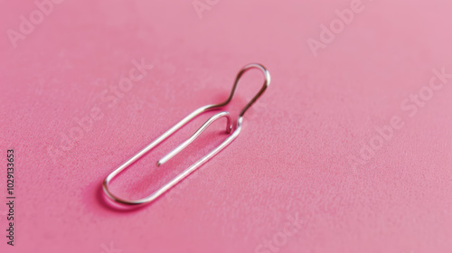 silver paperclip rests on soft pink background, showcasing its sleek design and functionality. This simple yet essential office supply adds touch of elegance to any workspace