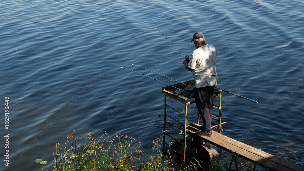 A fisherman on a platform with a cigarette in his teeth and fishing rods in his hands catches fish on a river or lake in the warm season. They stand with their backs to us. A fisherman on a platform with a cigarette in his teeth and fishing rods in his hands catches fish on a river or lake in the warm season. They stand with their backs to us.