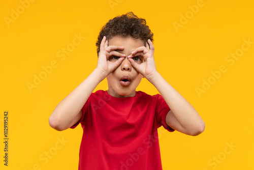 Studio portrait of cute little African American boy having fun making superhero's mask or imaginary binocular with his fingers, isolated over bright colored orange yellow background