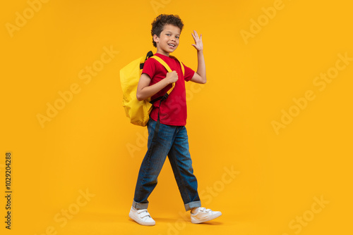 Full length studio portrait of cute casually dressed confident African American schoolboy walking with his bright colored backpack looking to the camera and waving hand, isolated over yellow backgroun