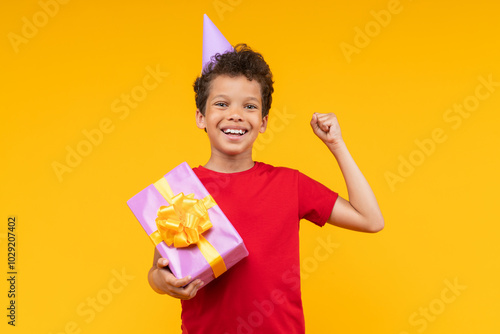 Studio portrait of cute happy smiling African American boy wearing plain red t-shirt and birthday hat posing over yellow background with decorated gift box in hand and clenching his fist