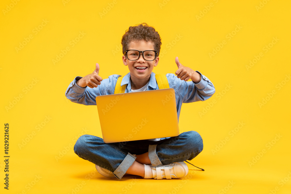 Cute happy smiling African American schoolboy in casual outfit and trendy eyeglasses posing sitting on bright colored yellow background with his laptop and giving thumbs up gesture with both hands Cute happy smiling African American schoolboy in casual outfit and trendy eyeglasses posing sitting on bright colored yellow background with his laptop and giving thumbs up gesture with both hands