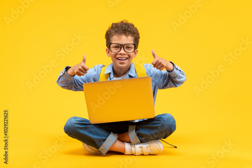 Cute happy smiling African American schoolboy in casual outfit and trendy eyeglasses posing sitting on bright colored yellow background with his laptop and giving thumbs up gesture with both hands