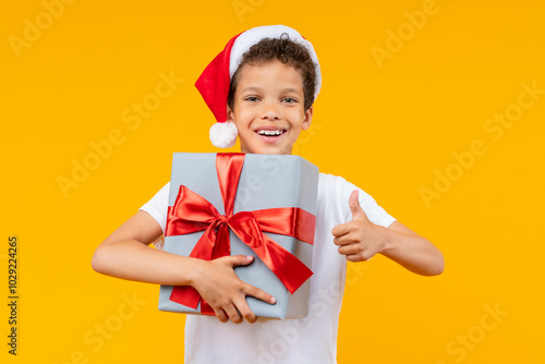 Studio shot of a joyful African American boy in Santa Claus hat posing over yellow background holding decorated gidt box in hand, smiling to the camera and making thumbs up gesture