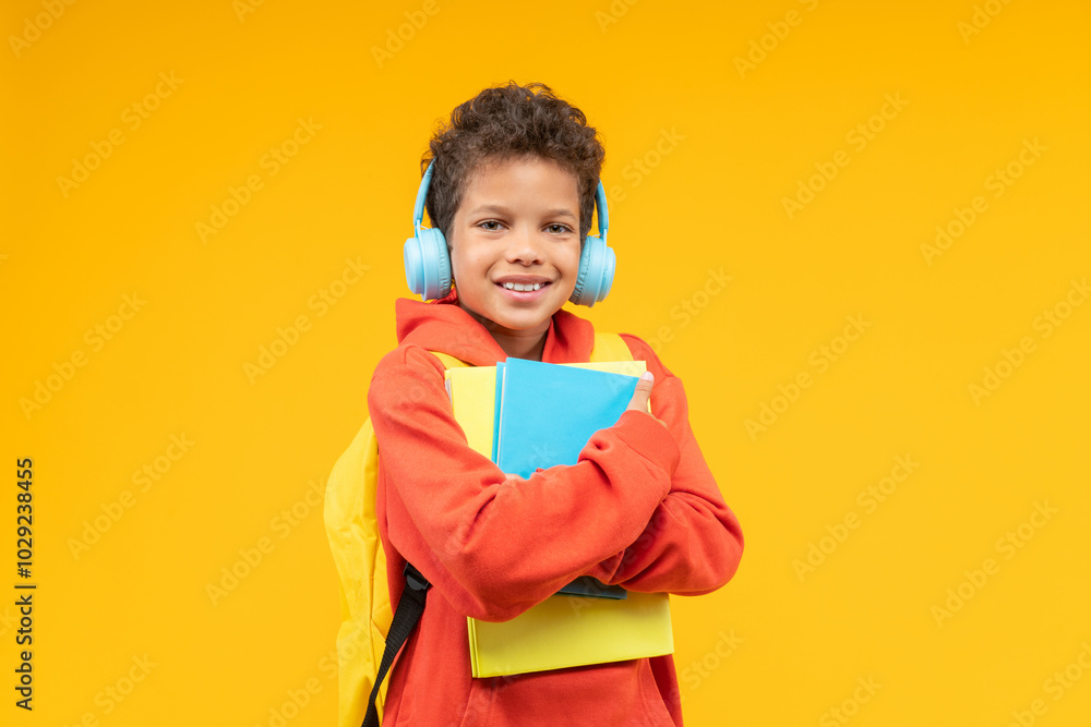 Studio shot of a cute smiling African American kid student wearing orange hoodie, bright backpack and wireless headphones, posing over yellow background with a pile of books in hands Studio shot of a cute smiling African American kid student wearing orange hoodie, bright backpack and wireless headphones, posing over yellow background with a pile of books in hands