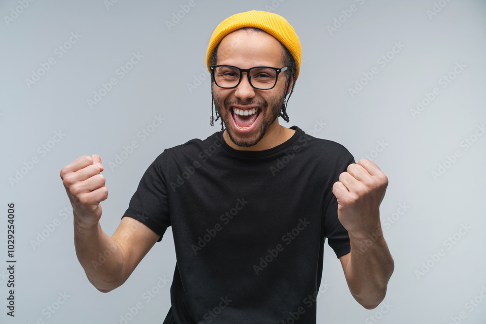 Studio portrait of joyful euphoric happy smiling stylish african american young man celebrating making winner's gesture clenching his fists shouting yes posing over plain grey background Studio portrait of joyful euphoric happy smiling stylish african american young man celebrating making winner's gesture clenching his fists shouting yes posing over plain grey background