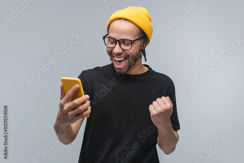 Studio portrait of stylish excited happy young African American man in trendy eyeglasses, basic black t-shirt and yellow beanie hat looking at his smartphone and making winner's gesture clenching fist
