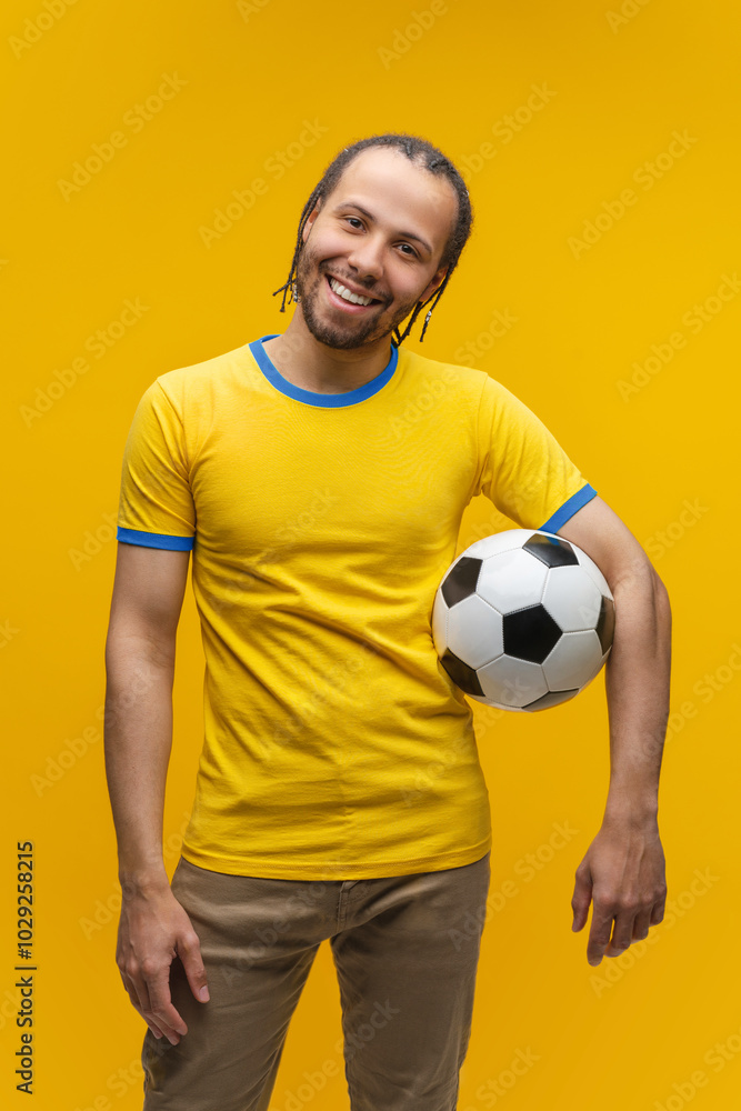 Vertical portrait of attractive football supporter with perfect broad smile wearing yellow blue t-shirt and posing with a soccer ball standing over bright colored yellow background Vertical portrait of attractive football supporter with perfect broad smile wearing yellow blue t-shirt and posing with a soccer ball standing over bright colored yellow background