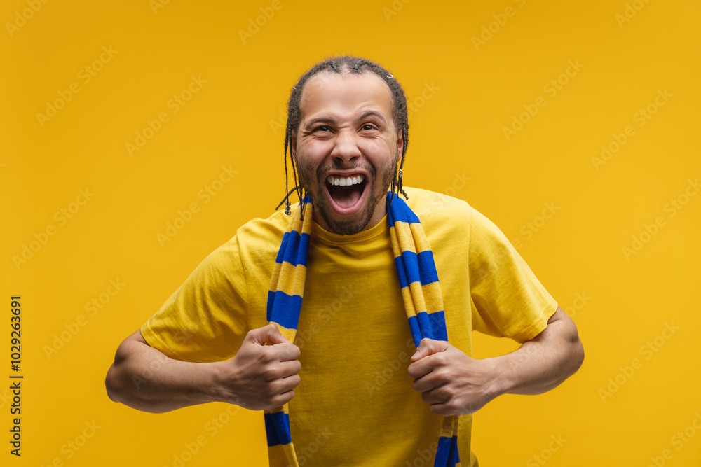 Studio shot of young euphoric African American male supporter wearing yellow blue scarf cheering for favorite team, screaming and celebrating with triumph, isolated on yellow background Studio shot of young euphoric African American male supporter wearing yellow blue scarf cheering for favorite team, screaming and celebrating with triumph, isolated on yellow background