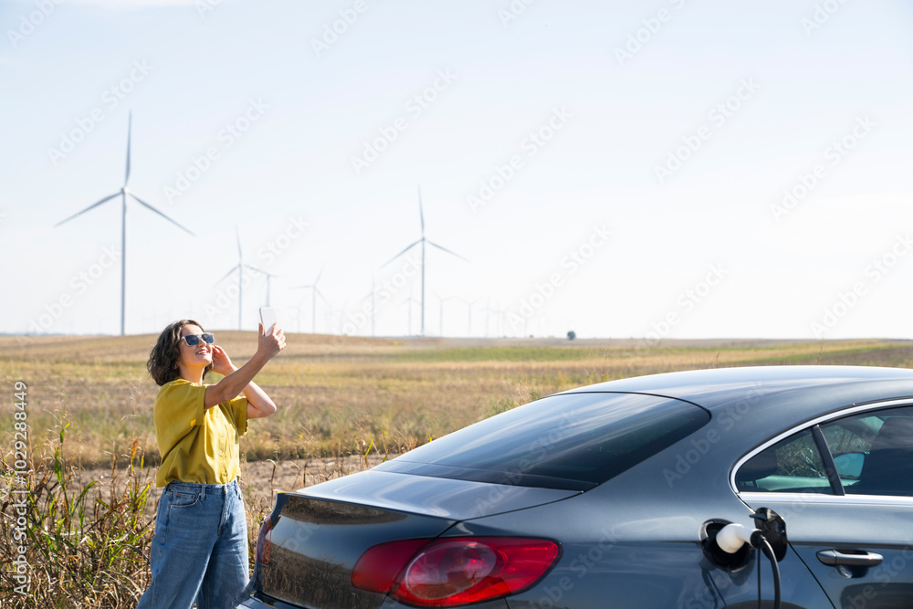 Woman with curve hairs wearing yellow shirt and sunglasses stands next to charging electric car and holds smartphone. Wind turbines in the background Woman with curve hairs wearing yellow shirt and sunglasses stands next to charging electric car and holds smartphone. Wind turbines in the background