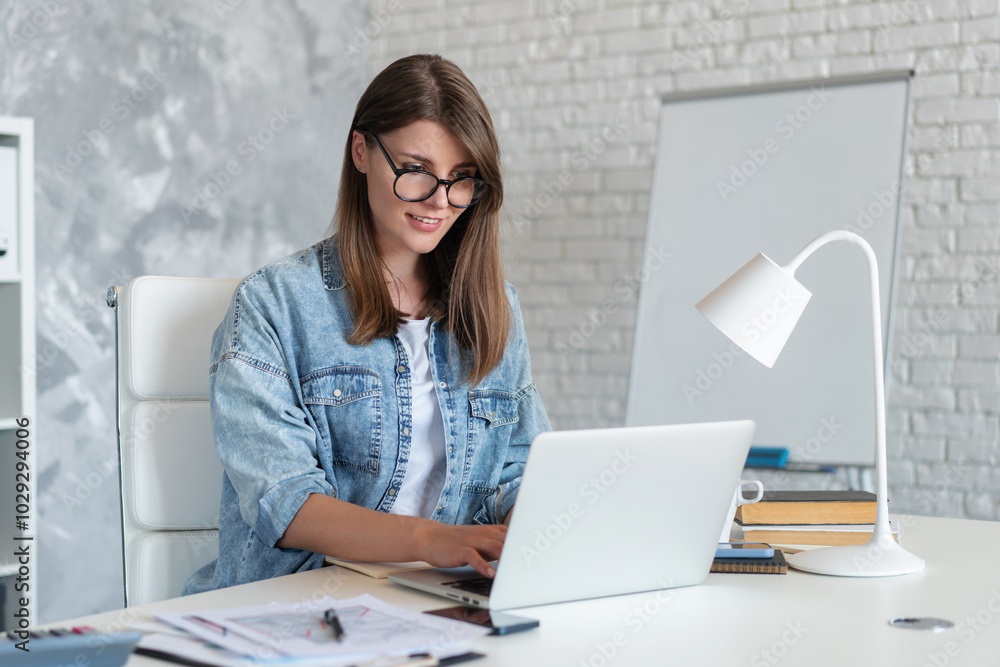 Portrait of casually dressed smiling pretty woman searching information on internet, working or studying, busy in self-education or ordering goods while sitting in light colored office or classroom Portrait of casually dressed smiling pretty woman searching information on internet, working or studying, busy in self-education or ordering goods while sitting in light colored office or classroom