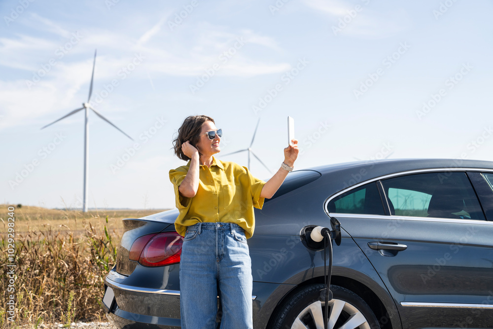Φωτογραφία Woman with curve hairs wearing yellow shirt and sunglasses stands next to charging electric car and holds smartphone