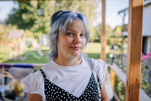 Young girl with blue hair, wearing a polka dot dress over a white t-shirt, standing outdoors on a sunny day with a relaxed expression.. photo