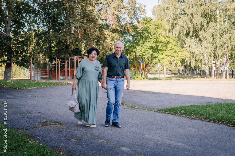An elderly couple walking hand in hand outdoors on a sunny day, enjoying each other’s company with a backdrop of trees and a playground.. An elderly couple walking hand in hand outdoors on a sunny day, enjoying each other’s company with a backdrop of trees and a playground..