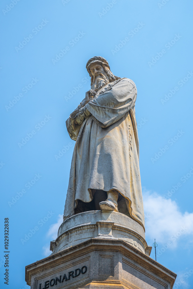 The monument showcases a detailed statue of Leonardo da Vinci, standing in a thoughtful pose atop a tall pedestal in Milan's historic Piazza della Scala, under a clear blue sky. The monument showcases a detailed statue of Leonardo da Vinci, standing in a thoughtful pose atop a tall pedestal in Milan's historic Piazza della Scala, under a clear blue sky.