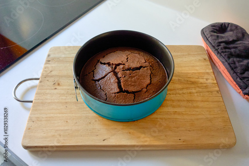 A freshly baked chocolate cake cracked in a round blue baking pan, placed on a wooden cutting board next to an induction stove and a black oven mitt, ready to be served