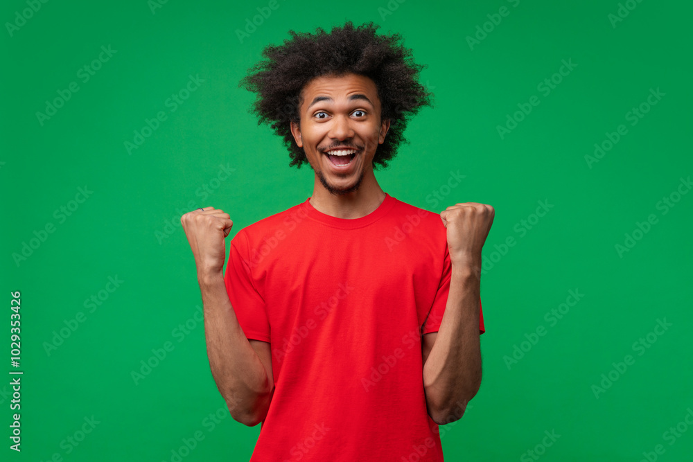 Fototapeta premium Studio portrait of joyful astonished happy smiling african american young man celebrating making winner's gesture clenching his fists with excited face expression posing over green background