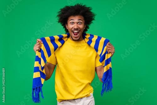 Portrait of excited happy smiling young african american soccer supporter man in basic t-shirt and yellow-blue scarf cheering for his favourite team