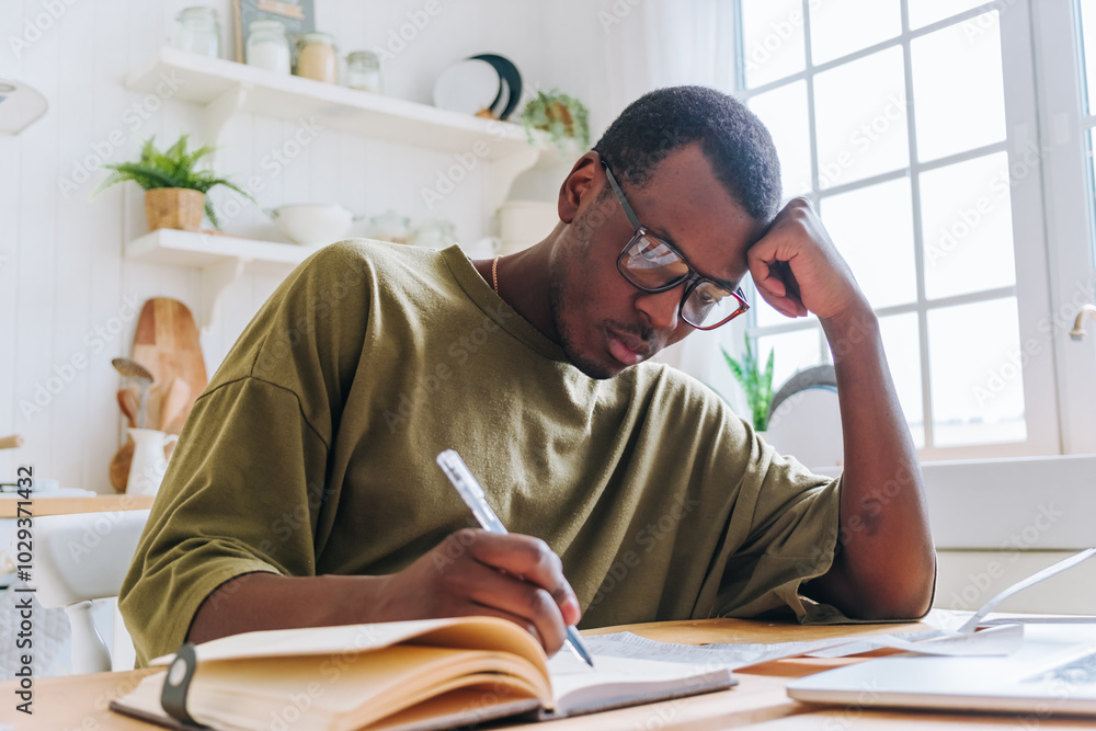 Young black man studying at home, focused on taking notes from a textbook, using a laptop for resources, surrounded by educational materials and natural light Young black man studying at home, focused on taking notes from a textbook, using a laptop for resources, surrounded by educational materials and natural light