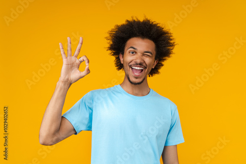 Portrait of young happy smiling African American man posing in studio over bright colored orange yellow background winking and showing ok sign gesture
