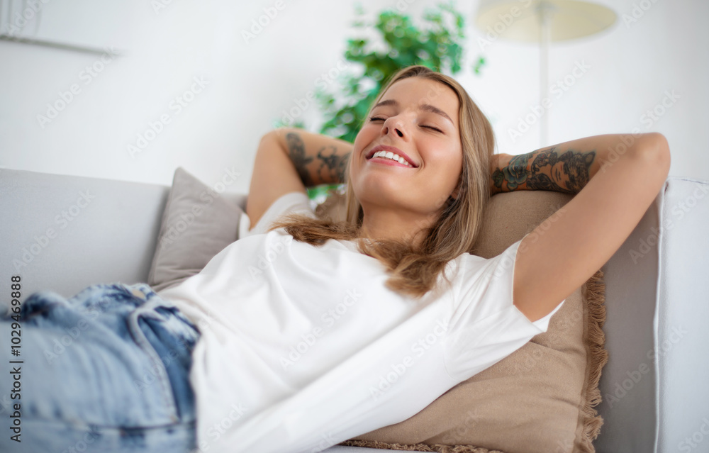 Beautiful blonde woman lounges back on a sofa with her arms behind her head, smiling joyfully. She wears a casual white t-shirt and jeans, relaxing in a bright, modern living room with soft pillows Beautiful blonde woman lounges back on a sofa with her arms behind her head, smiling joyfully. She wears a casual white t-shirt and jeans, relaxing in a bright, modern living room with soft pillows