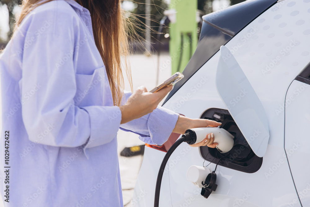Female Charging Electric Car And Holding Smartphone Outdoors Depicting Sustainable Transportation And Modern Technology Lifestyle Female Charging Electric Car And Holding Smartphone Outdoors Depicting Sustainable Transportation And Modern Technology Lifestyle