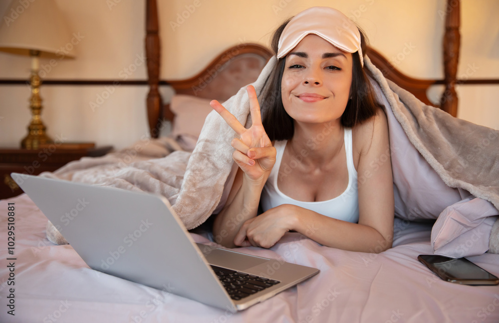 A smiling woman in bed, wearing a sleep mask, flashes a peace sign while using a laptop. A phone rests beside her. The cozy bedroom setting suggests relaxation, positivity, and modern comfort. A smiling woman in bed, wearing a sleep mask, flashes a peace sign while using a laptop. A phone rests beside her. The cozy bedroom setting suggests relaxation, positivity, and modern comfort.