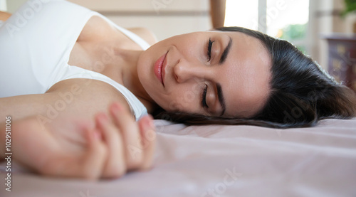 A close-up of a peaceful woman resting in bed, eyes closed, wearing a white tank top. The soft light highlights her serene expression, creating a calm and relaxed morning or nap time atmosphere. photo