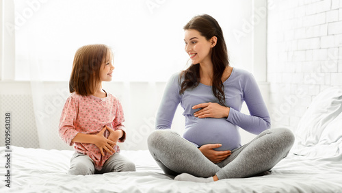 Expecting mother telling little girl about pregnancy, pregnant mother and preschool daughter touching bellies, sitting on bed