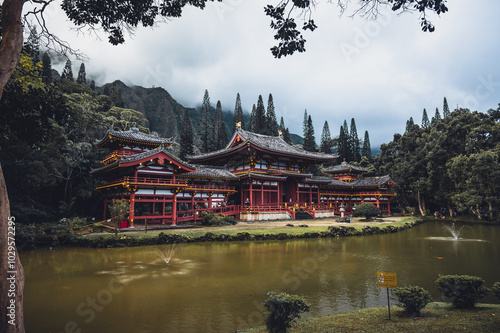 A tranquil view of an elegant traditional temple beside a serene pond, surrounded by mountains on a cloudy day