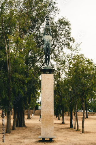 Statue of a warrior holding a torch atop a marble pedestal in a tranquil park surrounded by trees during the day