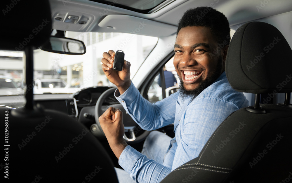 Own Car. Joyful African American Guy Holding Auto Key Shaking Fists Sitting In Automobile In Dealership Shop Own Car. Joyful African American Guy Holding Auto Key Shaking Fists Sitting In Automobile In Dealership Shop