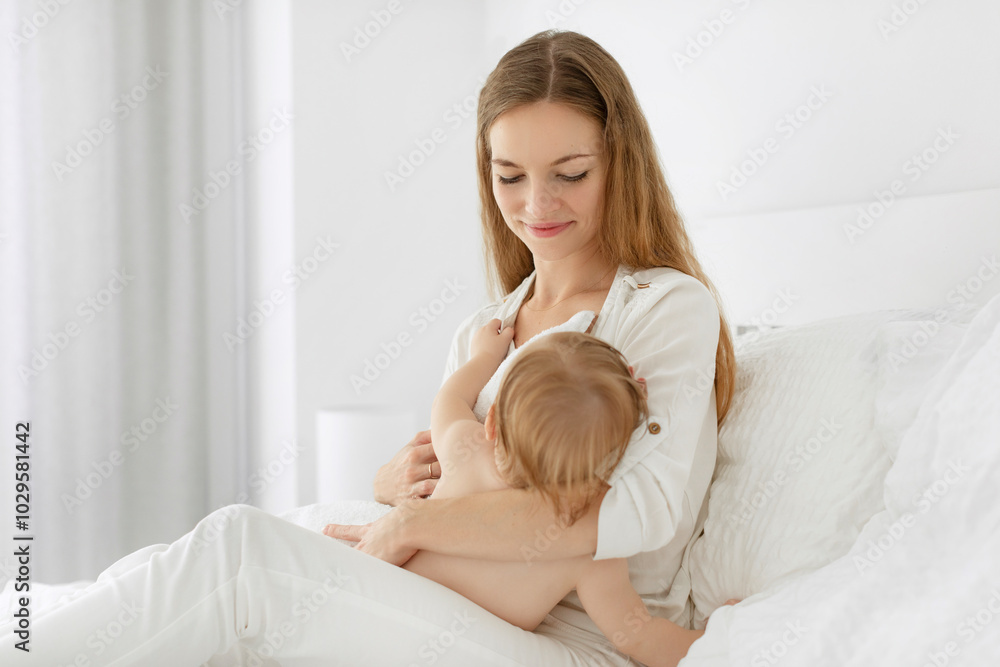 Beautiful mother breast-feeding her baby girl, sitting on bed and hugging little daughter, proper healthy breastfeeding of children Beautiful mother breast-feeding her baby girl, sitting on bed and hugging little daughter, proper healthy breastfeeding of children